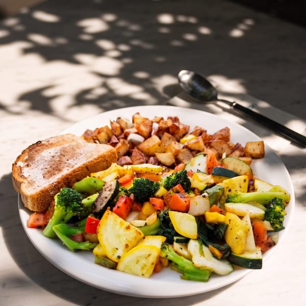 Plate of roasted mixed vegetables, potatoes, and sliced bread on a restaurant table with spoon and knife.