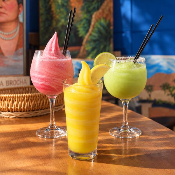 Three colorful frozen drinks sit on a sunlit wooden table: pink, yellow, and green beverages garnished with fruit and straws.